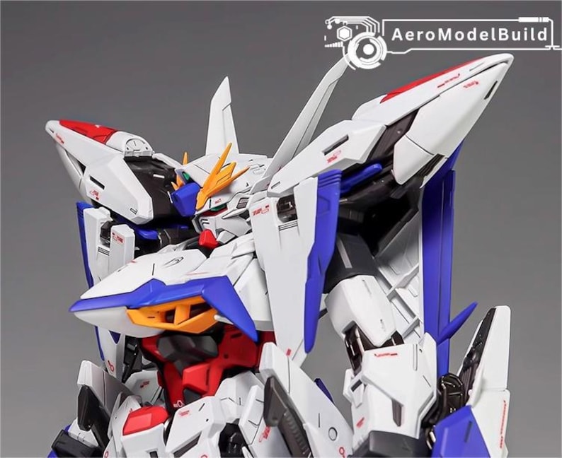 Puede incluir: Primer plano de un modelo de robot blanco, azul, rojo y amarillo. El modelo tiene detalles intrincados, &aacute;ngulos agudos y un dise&ntilde;o futurista. El texto "AeroModelBuild" es visible en la esquina superior derecha.