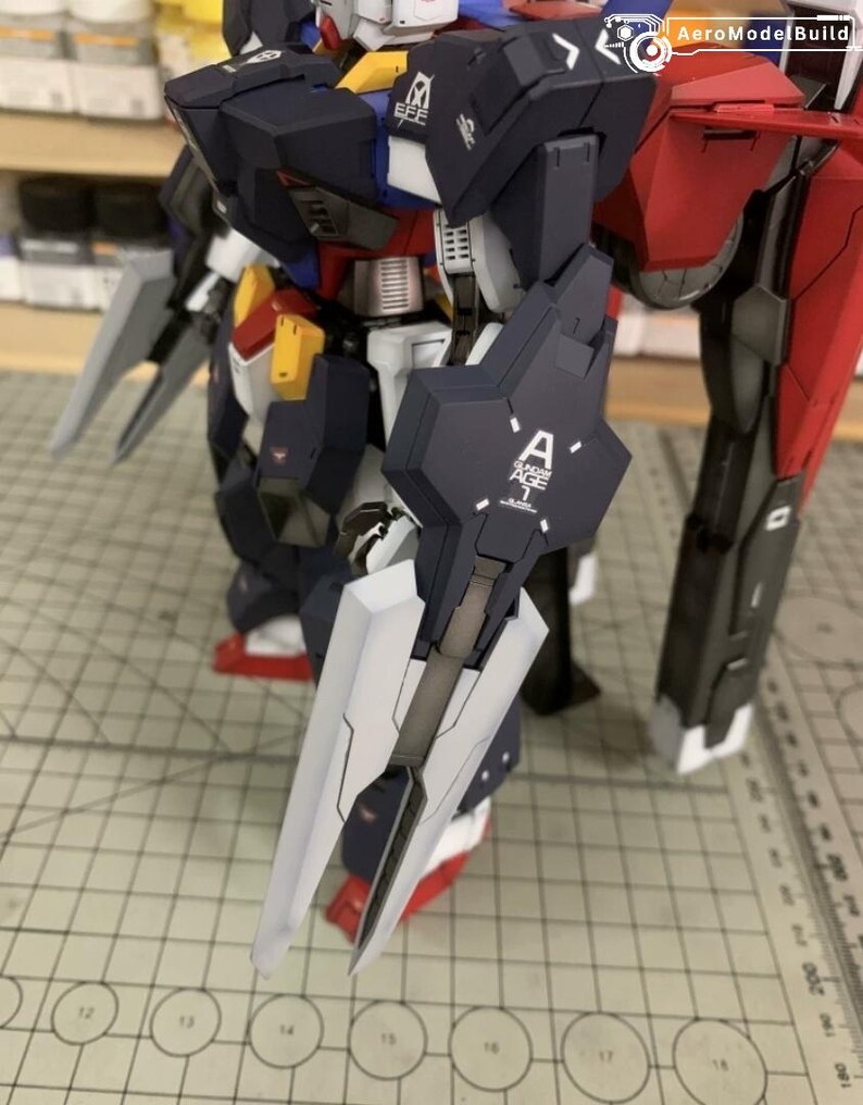 Puede incluir: Modelo detallado de un robot con esquema de color azul oscuro y rojo. El robot tiene un escudo grande con la letra "A" y el texto "AGE 1" impreso. El modelo est&aacute; sobre una superficie cuadriculada.