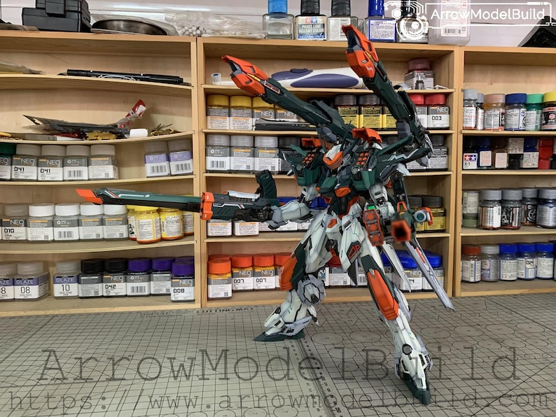 May include: A detailed model robot with a complex design, featuring white, orange, and green armor. The robot is posed with a weapon and wings, set against a backdrop of shelves filled with model-building supplies. The text "ArrowModelBuild" is visible.