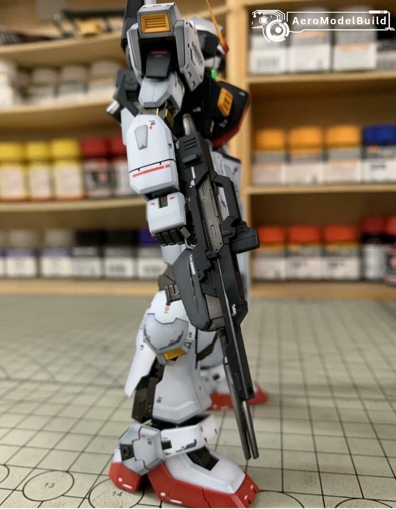 Pode incluir: Um modelo detalhado de um rob&ocirc; branco, preto e cinza com detalhes vermelhos, segurando um rifle preto. O modelo tem detalhes intrincados e est&aacute; posicionado em uma superf&iacute;cie quadriculada. O logotipo AeroModelBuild &eacute; vis&iacute;vel no fundo.