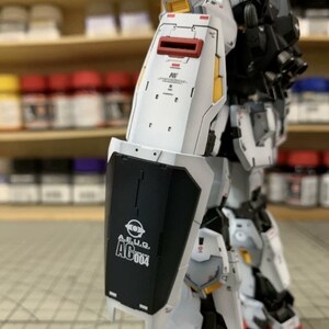 Pode incluir: Modelo de rob&ocirc; branco e preto parcialmente montado com detalhes em vermelho e amarelo. O modelo apresenta um escudo com o texto "A.E.U.G. AG004". O fundo mostra prateleiras com potes de tinta.
