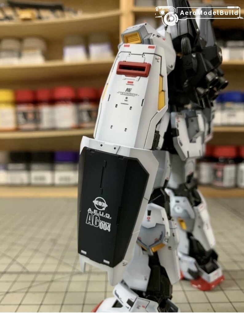 Pode incluir: Modelo de rob&ocirc; branco e preto parcialmente montado com detalhes em vermelho e amarelo. O modelo apresenta um escudo com o texto "A.E.U.G. AG004". O fundo mostra prateleiras com potes de tinta.