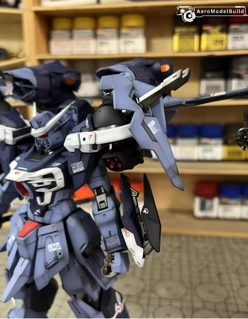May include: A detailed, partially assembled model kit of a futuristic robot, primarily in shades of blue and gray with red and white accents. The model features intricate mechanical details, including articulated limbs and various weapon-like attachments. The AeroModelBuild logo is visible.