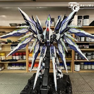 May include: A highly detailed model of a futuristic aircraft or robot, with elaborate wing-like features in white, purple, and blue. The model is mounted on a black base, highlighting the complex design. Shelves in the background display paint supplies.