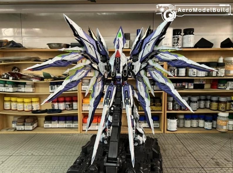 May include: A highly detailed model of a futuristic aircraft or robot, with elaborate wing-like features in white, purple, and blue. The model is mounted on a black base, highlighting the complex design. Shelves in the background display paint supplies.
