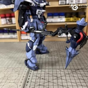 May include: A detailed model of a robot warrior, primarily in shades of blue and gray, holding a large weapon. The model is standing on a grid-patterned surface, with shelves of paint bottles in the background. The robot has intricate details.