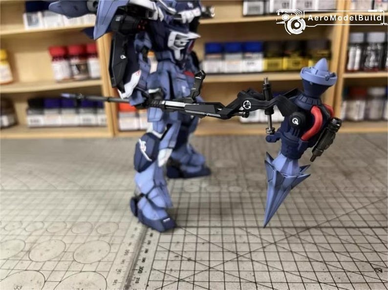 May include: A detailed model of a robot warrior, primarily in shades of blue and gray, holding a large weapon. The model is standing on a grid-patterned surface, with shelves of paint bottles in the background. The robot has intricate details.