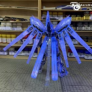 May include: A clear blue model kit of a futuristic robot with large, wing-like structures extending from its back. The model is partially assembled and stands on a grey surface, with shelves of model supplies in the background.