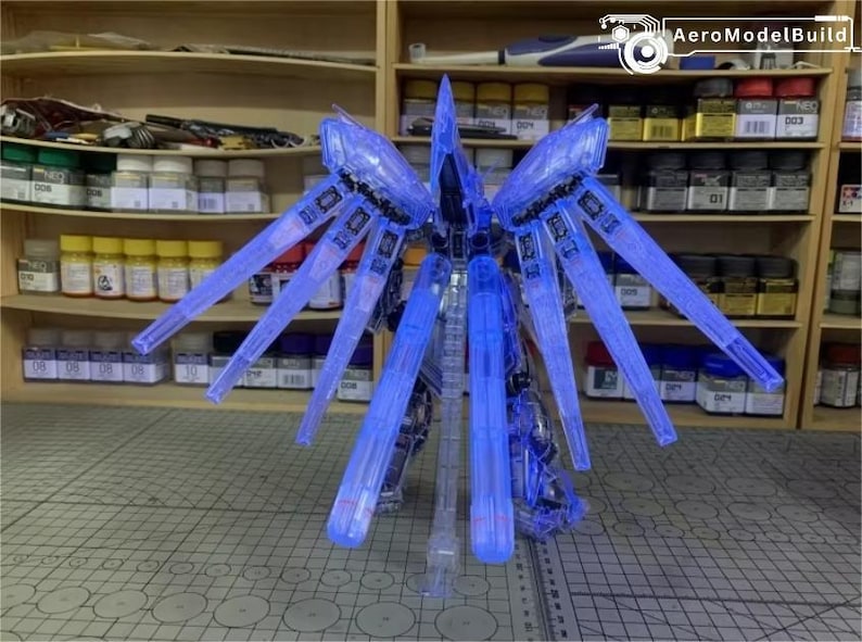 May include: A clear blue model kit of a futuristic robot with large, wing-like structures extending from its back. The model is partially assembled and stands on a grey surface, with shelves of model supplies in the background.