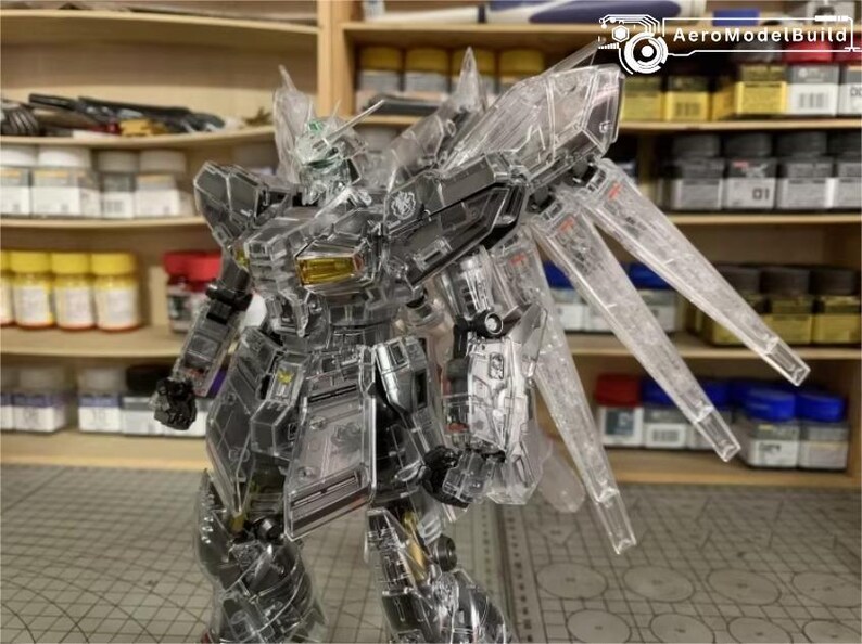May include: A transparent plastic robot model with articulated limbs and wings. The robot features a detailed design with visible internal components. The model is posed dynamically, highlighting its intricate construction. The background includes shelves with paint supplies.