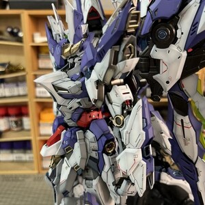 May include: A highly detailed model robot, predominantly white, purple, red, and gold. The design incorporates intricate mechanical elements and sharp angles, giving a futuristic appearance. The background is blurred, focusing attention on the model's complex structure.