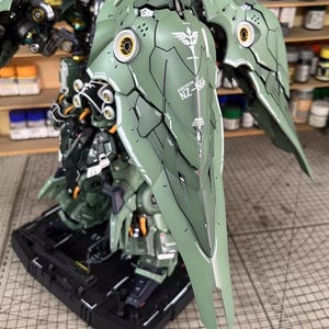 May include: A detailed model of a green and white robot with large wing-like structures. The model features intricate paneling, gold accents, and the text "NZ-666". It is mounted on a black base, showcasing a complex design.