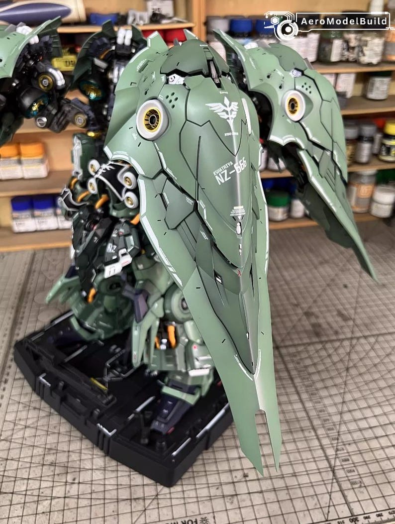 May include: A detailed model of a green and white robot with large wing-like structures. The model features intricate paneling, gold accents, and the text "NZ-666". It is mounted on a black base, showcasing a complex design.