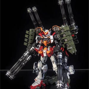 May include: A detailed model of a Gundam robot, with a colour scheme of white, red, orange, and green. The robot is heavily armed with multiple large cannons and missile launchers, showcasing a complex design. The AeroModelBuild logo is visible.