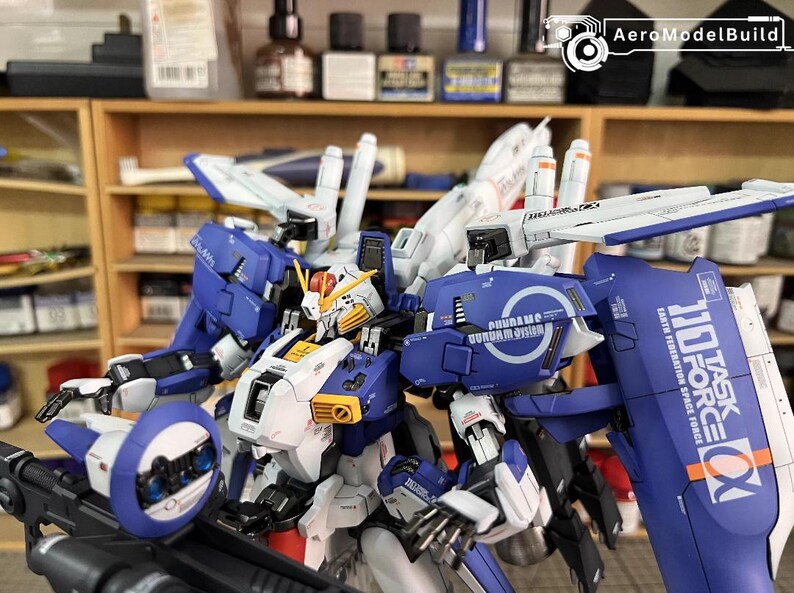 May include: A detailed model of a Gundam robot, mainly blue and white, with yellow and red details. The model features multiple panels, wings, and a shield with text. The image includes the text "AeroModelBuild".