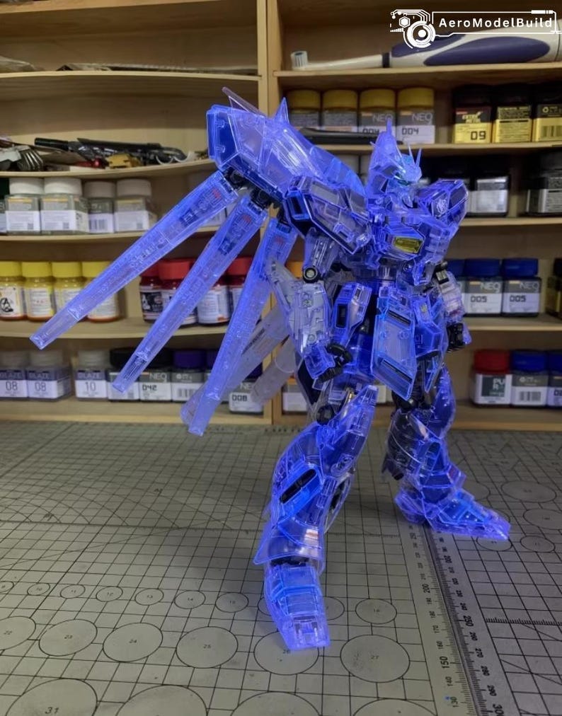 May include: A transparent blue model robot with illuminated wings and body parts. The robot is standing on a gray surface with a ruler. The background includes shelves with various containers and the AeroModelBuild logo.