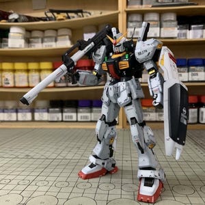 Pode incluir: Modelo detalhado de um rob&ocirc; Gundam, predominantemente branco com detalhes em preto, vermelho e amarelo. O rob&ocirc; est&aacute; segurando um grande rifle branco e preto e tem um escudo no bra&ccedil;o esquerdo. O modelo est&aacute; em uma superf&iacute;cie quadriculada.