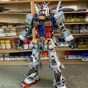 K&ouml;nnte beinhalten: Ein detailliertes Modell eines Gundam-Roboters mit einer Farbgebung aus Wei&szlig;, Blau, Rot und Gelb. Der Roboter steht mit leicht ausgestreckten Armen. Das Modell steht auf einer Schneidematte, im Hintergrund Regale mit Farbe und Werkzeugen.
