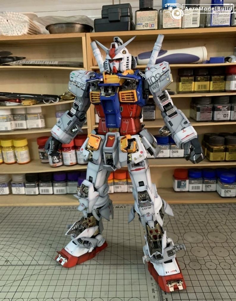 K&ouml;nnte beinhalten: Ein detailliertes Modell eines Gundam-Roboters mit einer Farbgebung aus Wei&szlig;, Blau, Rot und Gelb. Der Roboter steht mit leicht ausgestreckten Armen. Das Modell steht auf einer Schneidematte, im Hintergrund Regale mit Farbe und Werkzeugen.