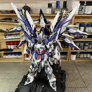 May include: A highly detailed model robot, predominantly white, purple, and blue, with elaborate wing-like structures. The model features intricate detailing and stands on a black base. The design suggests a futuristic aesthetic.