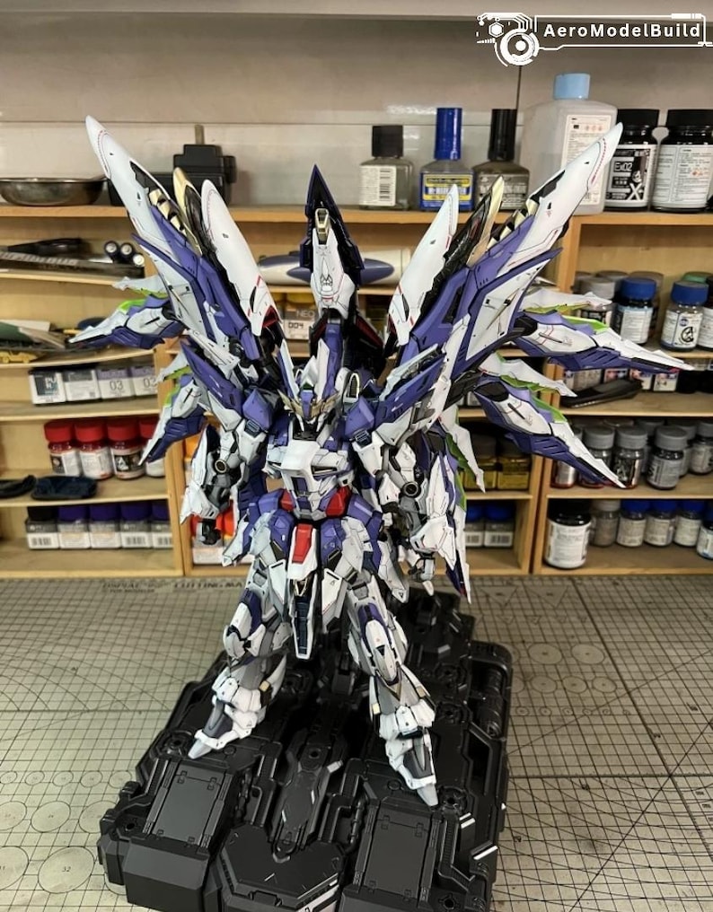 May include: A highly detailed model robot, predominantly white, purple, and blue, with elaborate wing-like structures. The model features intricate detailing and stands on a black base. The design suggests a futuristic aesthetic.