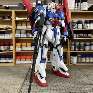 May include: A detailed model of a robot with a white, blue, red, and yellow color scheme. The robot is standing on a gray surface with a ruler. The robot has a large gun and red wings. The background includes shelves with paint bottles and the text "ArrowModelBuild".