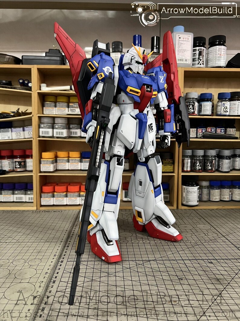 May include: A detailed model of a robot with a white, blue, red, and yellow color scheme. The robot is standing on a gray surface with a ruler. The robot has a large gun and red wings. The background includes shelves with paint bottles and the text "ArrowModelBuild".