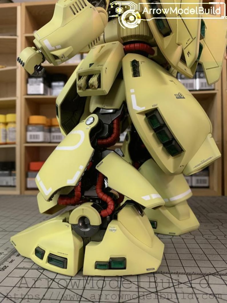 May include: A detailed, close-up view of a yellow model robot's lower body. The model features articulated legs, with red tubing and white accent lines. The image includes the text "ArrowModelBuild" in the upper right corner.