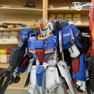 May include: A detailed model kit of a Gundam robot, with a blue, white, red, and orange colour scheme. The robot is posed with a large weapon and a red fin on its back. The model is set against a backdrop of shelves with various containers.