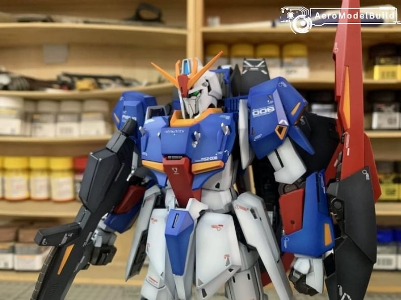 May include: A detailed model kit of a Gundam robot, with a blue, white, red, and orange colour scheme. The robot is posed with a large weapon and a red fin on its back. The model is set against a backdrop of shelves with various containers.