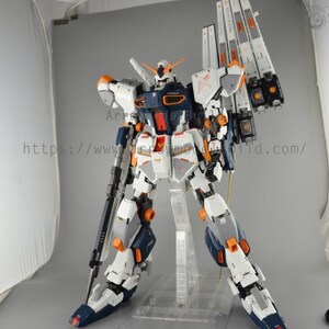 May include: A detailed model of a robot, primarily white, navy blue, and orange. The robot has a complex design with multiple panels, joints, and weapons. It stands on a clear plastic stand against a neutral background. The model is a collectible item.