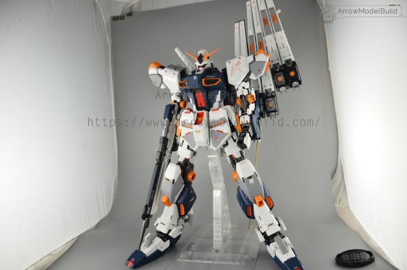 May include: A detailed model of a robot, primarily white, navy blue, and orange. The robot has a complex design with multiple panels, joints, and weapons. It stands on a clear plastic stand against a neutral background. The model is a collectible item.