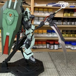 Puede incluir: Maqueta detallada de un robot Gundam, con una combinaci&oacute;n de colores verde y blanco, un gran escudo y una larga hoja curva. El modelo est&aacute; montado sobre un soporte negro, mostrando intrincados detalles mec&aacute;nicos.