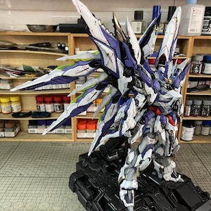 May include: A highly detailed mecha robot model, featuring white, purple, and blue colour scheme, with elaborate wing-like elements. The model is displayed on a black base, highlighting its intricate design. The AeroModelBuild logo is visible in the top right corner.