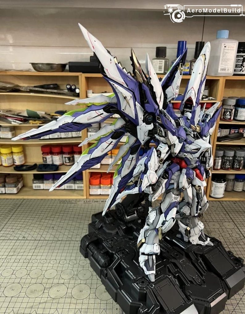 May include: A highly detailed mecha robot model, featuring white, purple, and blue colour scheme, with elaborate wing-like elements. The model is displayed on a black base, highlighting its intricate design. The AeroModelBuild logo is visible in the top right corner.