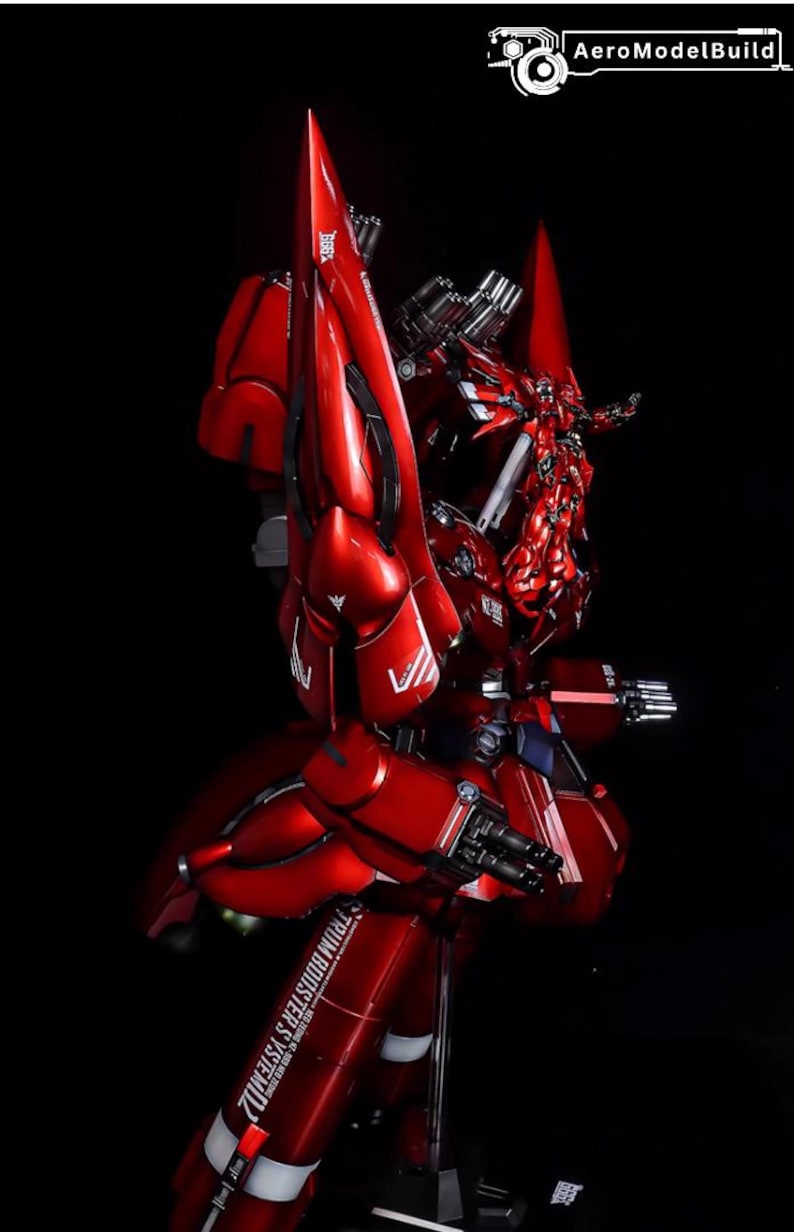 May include: A detailed, red model of a mobile suit robot, with intricate mechanical details and sharp angles. The model features multiple weapons and boosters, set against a black background. The text "TRUM BOOSTER'S SYSTEM" is visible.