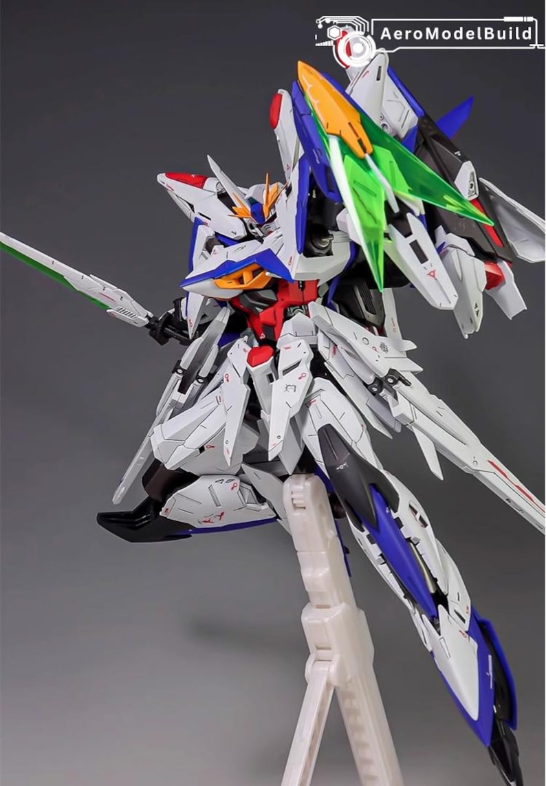 Puede incluir: Modelo detallado de un robot futurista, predominantemente blanco con detalles azules, rojos y amarillos. Cuenta con grandes alas, una espada y est&aacute; montado sobre un soporte. El logotipo de AeroModelBuild es visible.