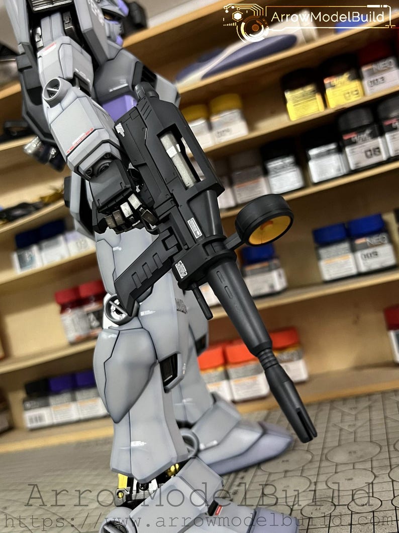 May include: A detailed, gray and black model robot figure holding a black weapon. The figure has articulated joints and is posed against a backdrop of paint supplies. The image includes the text "Arrow ModelBuild" and a website address.