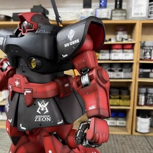 May include: A detailed model of a red and black robot with the text "MS-05RS" and "PRINCIPALITY OF ZEON". The robot has intricate mechanical details and is set against a blurred background of shelves with paint supplies.