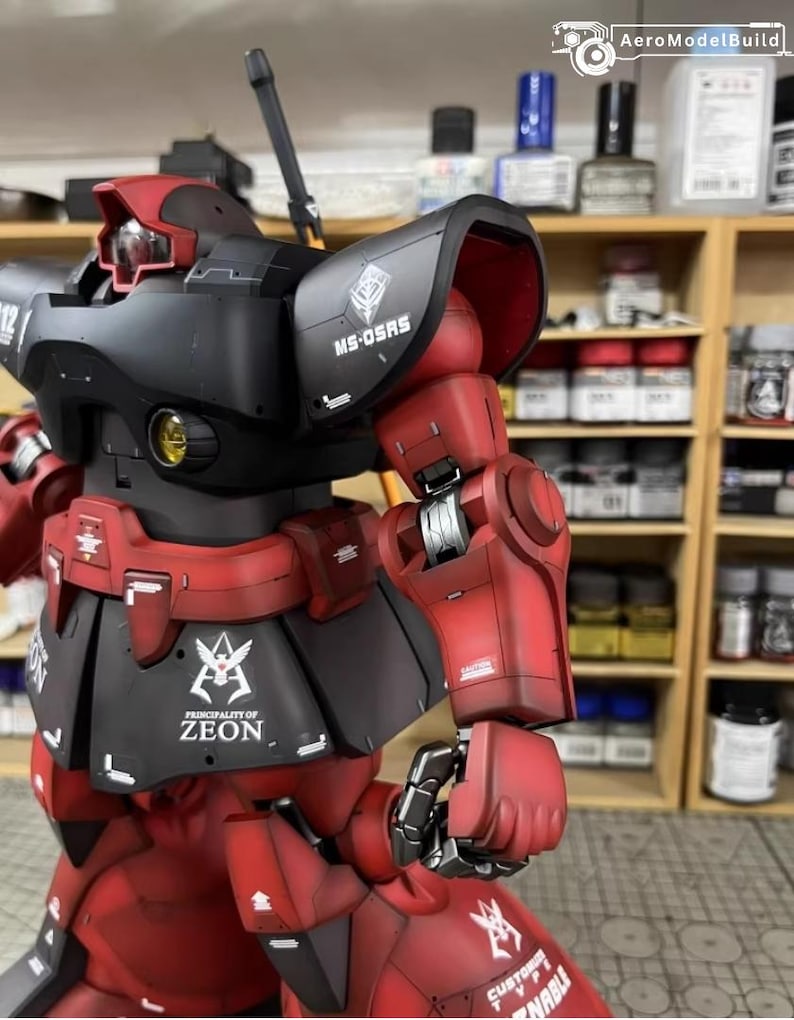 May include: A detailed model of a red and black robot with the text "MS-05RS" and "PRINCIPALITY OF ZEON". The robot has intricate mechanical details and is set against a blurred background of shelves with paint supplies.