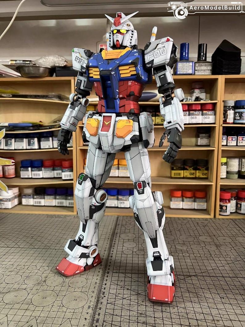 May include: A detailed model of a Gundam robot, featuring a color scheme of white, blue, red, yellow, and gray. The model has intricate panel lines and markings, with visible mechanical details. The robot is standing upright, showcasing its articulated limbs and weaponry.
