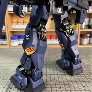 Pu&ograve; includere: Primo piano delle gambe e del busto inferiore di un modello di robot blu scuro e nero. Il modello presenta dettagli dorati e arancioni, con il numero "01" visibile. L'immagine mette in risalto gli intricati dettagli del modello.