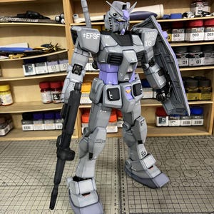 May include: A detailed model of a Gundam robot, primarily gray with purple and black accents. The robot is holding a black rifle and a shield. The model has the text "EFSF" and "WB 103" printed on it.