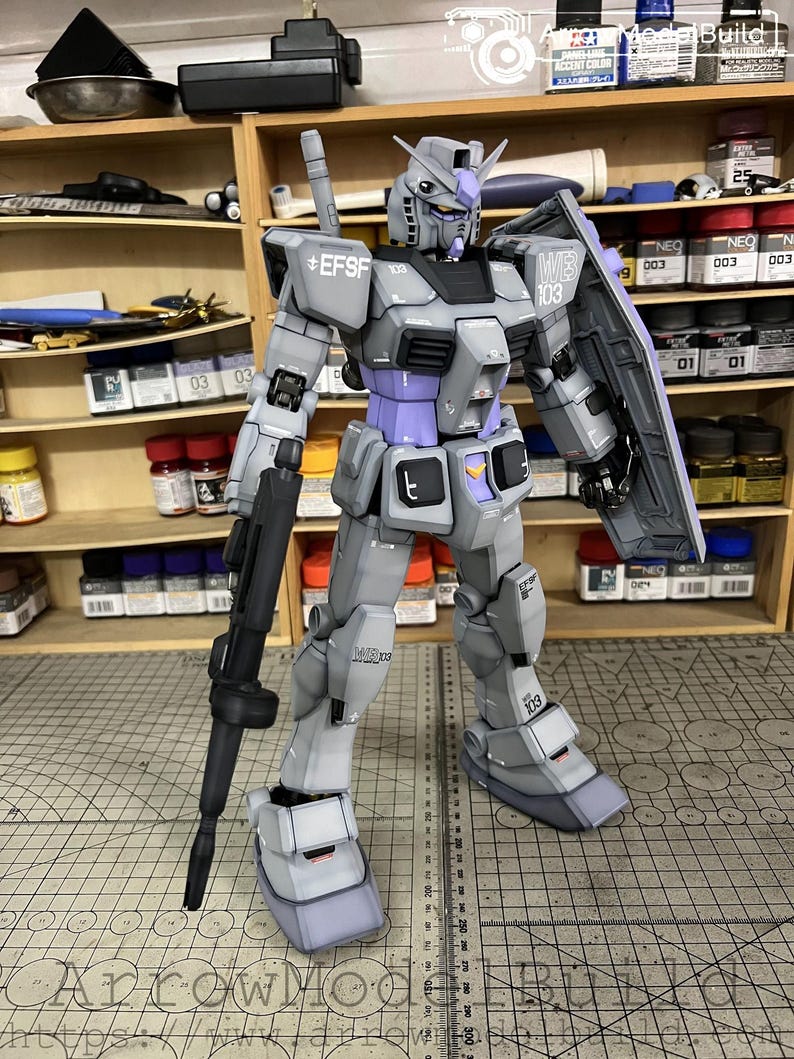 May include: A detailed model of a Gundam robot, primarily gray with purple and black accents. The robot is holding a black rifle and a shield. The model has the text "EFSF" and "WB 103" printed on it.