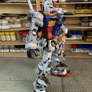 K&ouml;nnte beinhalten: Detailliertes Modell eines Gundam-Roboters mit einer Farbgebung aus Wei&szlig;, Blau, Rot und Gelb. Das Modell weist komplizierte mechanische Details auf, darunter eine gro&szlig;e Waffe auf dem R&uuml;cken. Das Modell steht auf einer grauen Oberfl&auml;che.