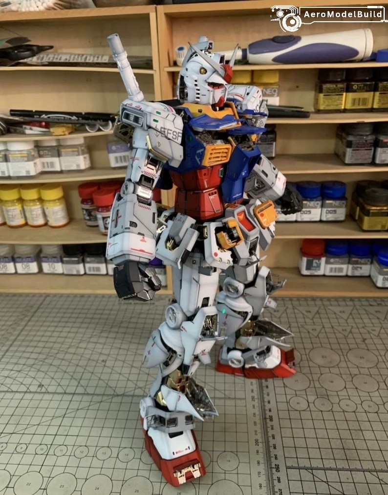 K&ouml;nnte beinhalten: Detailliertes Modell eines Gundam-Roboters mit einer Farbgebung aus Wei&szlig;, Blau, Rot und Gelb. Das Modell weist komplizierte mechanische Details auf, darunter eine gro&szlig;e Waffe auf dem R&uuml;cken. Das Modell steht auf einer grauen Oberfl&auml;che.