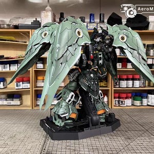 May include: A detailed model of a mech robot with large, articulated wings. The robot is primarily green and black with orange accents. The wings are a light green with intricate detailing. The model is on a black base, with the text "AeroModelBuild" visible.
