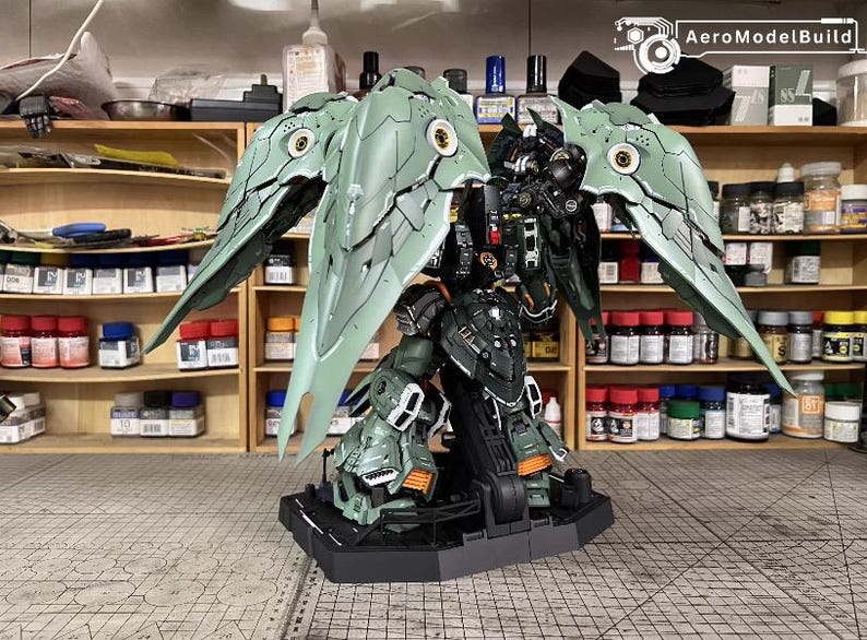 May include: A detailed model of a mech robot with large, articulated wings. The robot is primarily green and black with orange accents. The wings are a light green with intricate detailing. The model is on a black base, with the text "AeroModelBuild" visible.