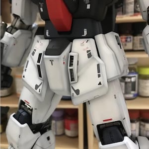 Op de afbeelding: Close-up van een gedetailleerde witte en zwarte modelrobot, met rode en oranje accenten. Het model heeft ingewikkelde paneellijnen en markeringen, wat wijst op een hoog niveau van vakmanschap. De afbeelding benadrukt de texturen en details van het model.