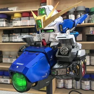 Könnte beinhalten: Ein detailliertes Modell eines Gundam-Roboters mit weißem, blauem und goldenem Farbschema. Der Roboter weist komplizierte mechanische Details auf, darunter ein grün leuchtendes Visier und eine große grüne Kugel mit dem Wort "ZERO". Das Modell wird auf einem schwarzen Ständer präsentiert.